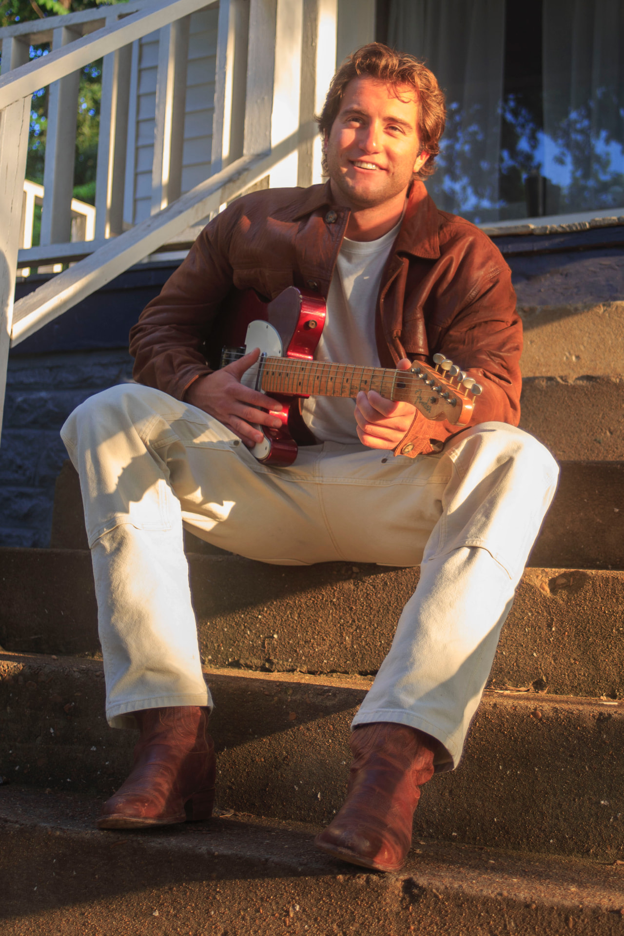 Dante DeRosa smiling on porch steps at golden hour holding his red Telecaster