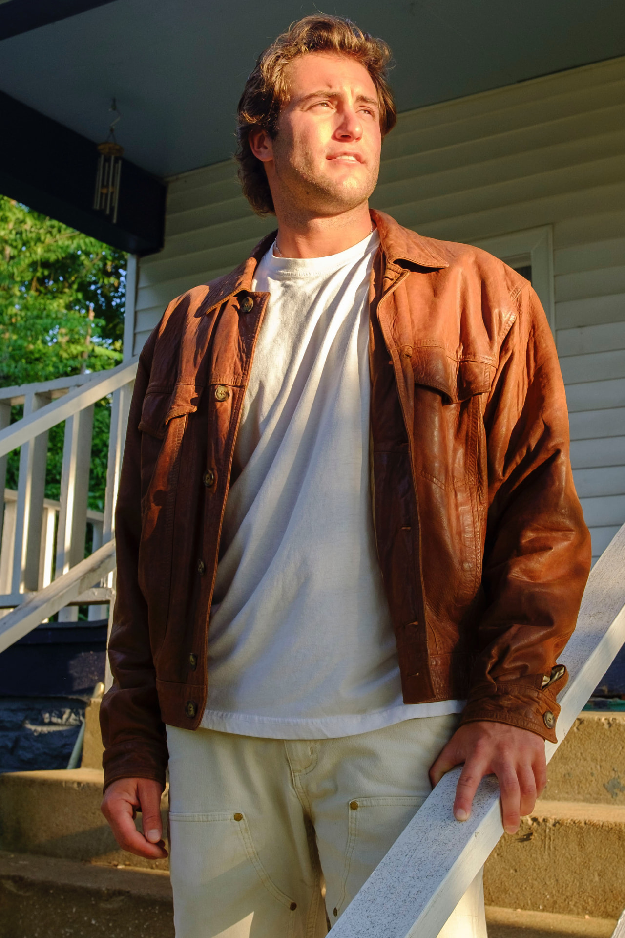 Dante DeRosa standing on a porch steps in a worn leather jacket, lit by warm evening sun